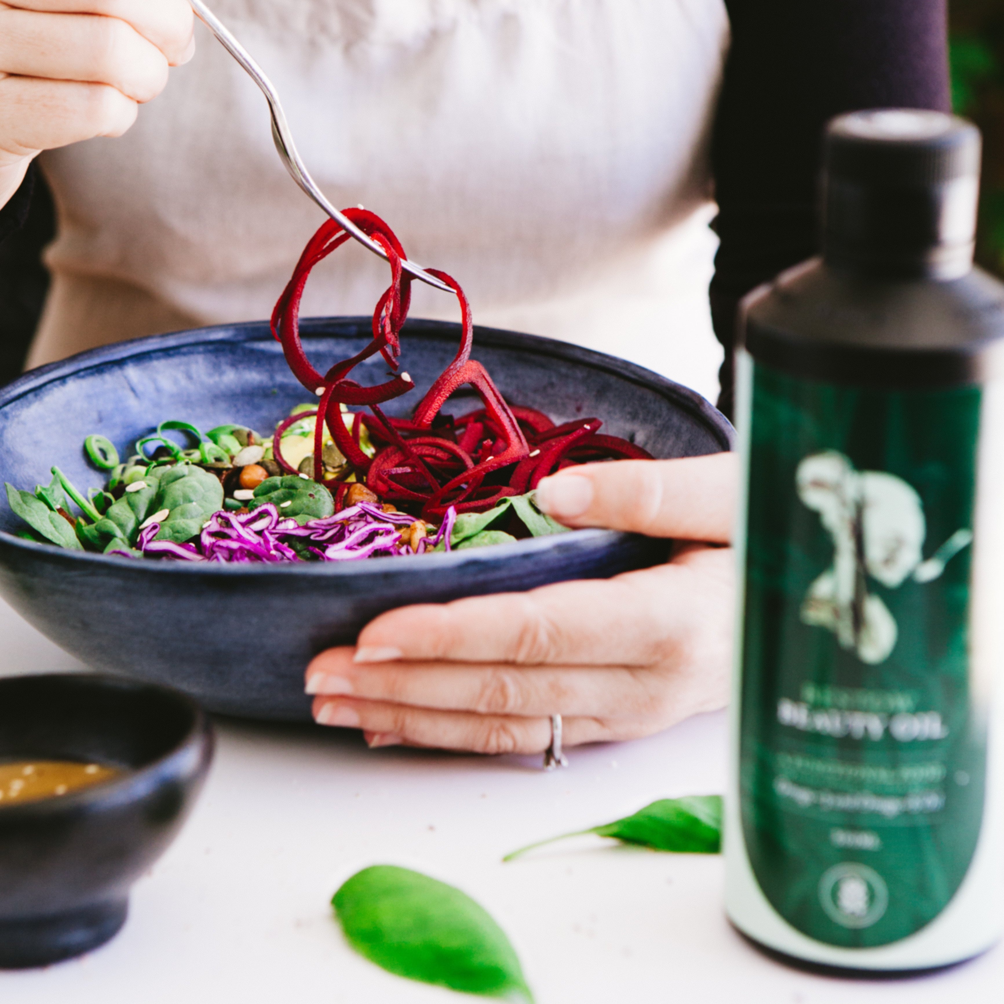 Person holding a bowl of salad with a bottle of Bestow Beauty oil on a table.