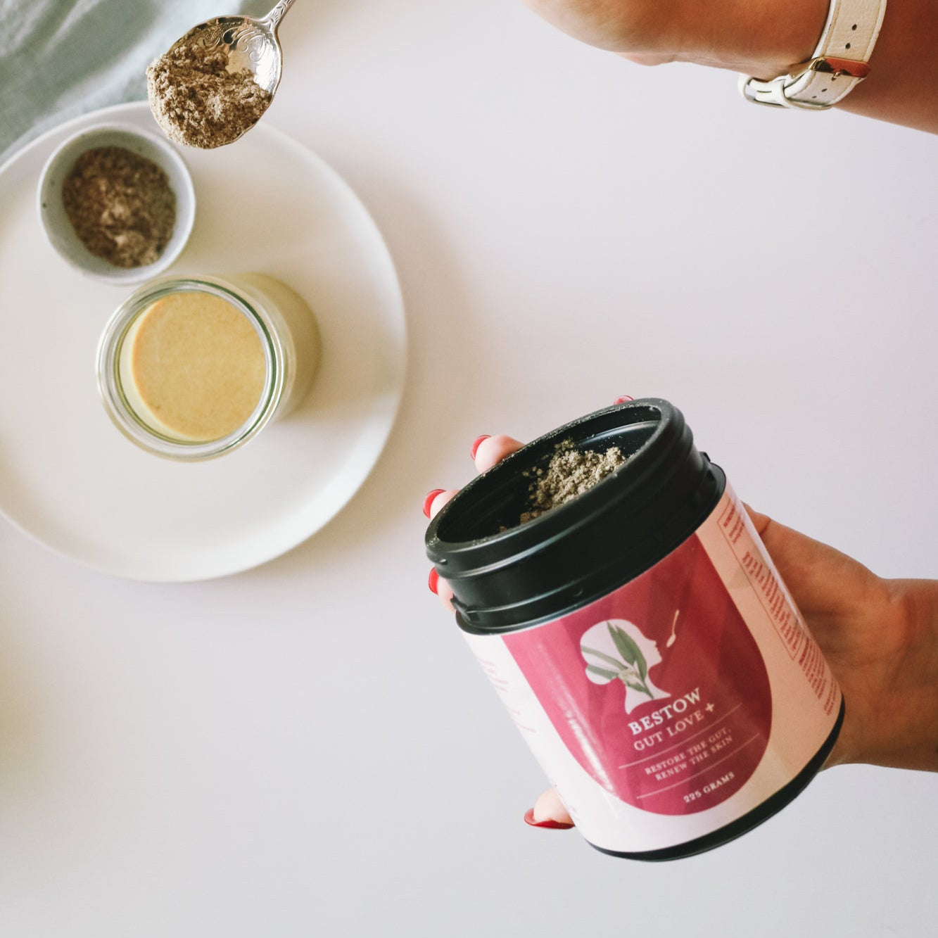 Person holding a container of Bestow Gut Love+ product with a spoonful of powder on a white surface.