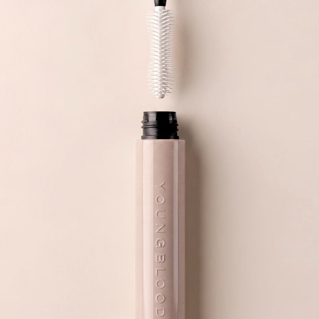 A close-up of a cosmetic tube in a pale blush pink color with a black cap, shown with its applicator wand pulled out above the open top. The brand name 'YOUNG BLOOD' is vertically embossed on the tube.