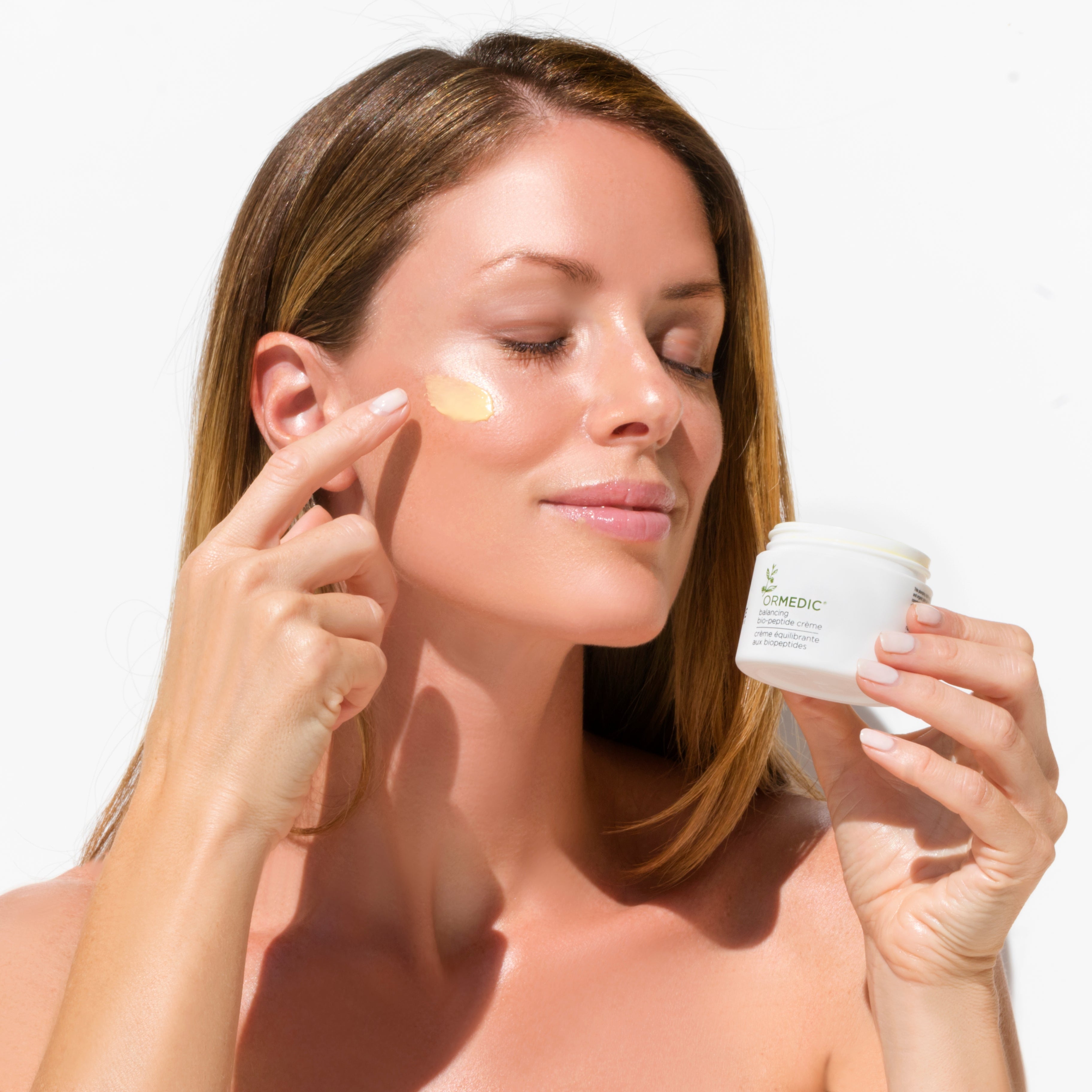 Woman applying cream to her face with a jar of cream in her hand on a white background