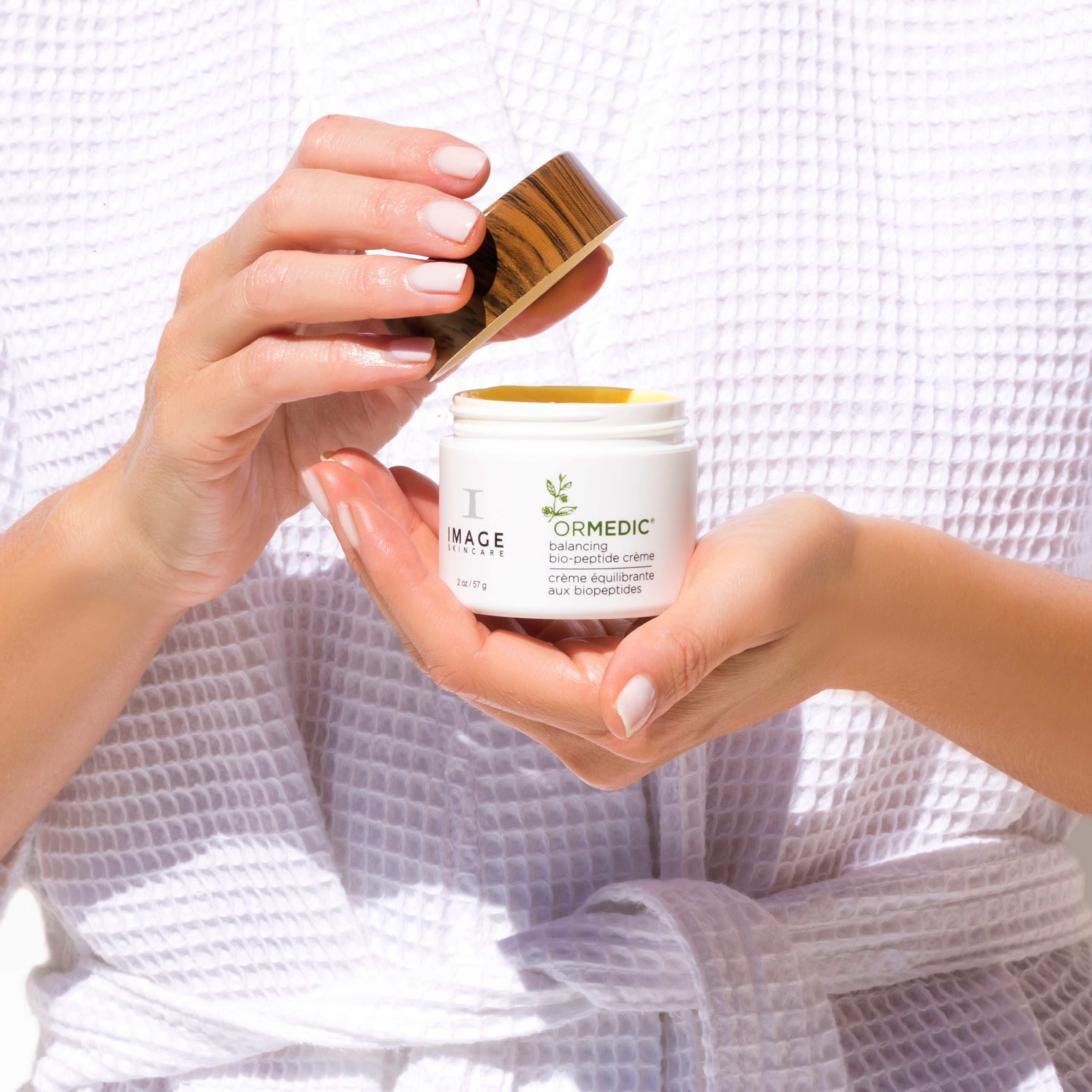 Person holding a jar of cream with a wooden lid, wearing a white robe.