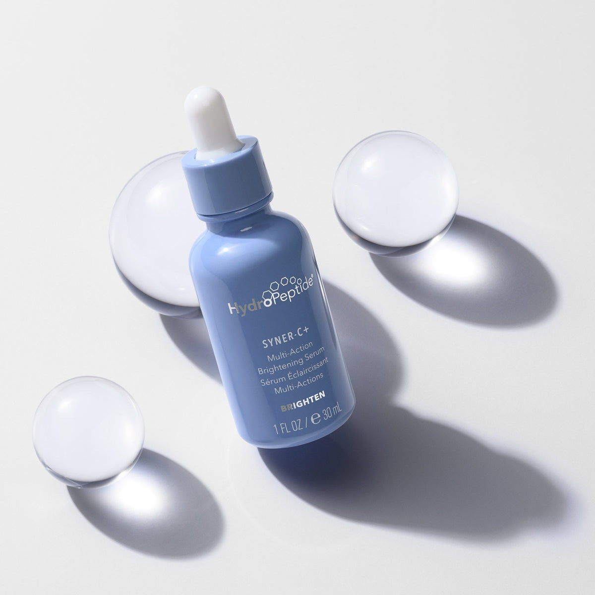 A light blue dropper bottle labeled HydroPeptide Syner-C+ on a clean white background, surrounded by four translucent spherical props casting soft shadows.