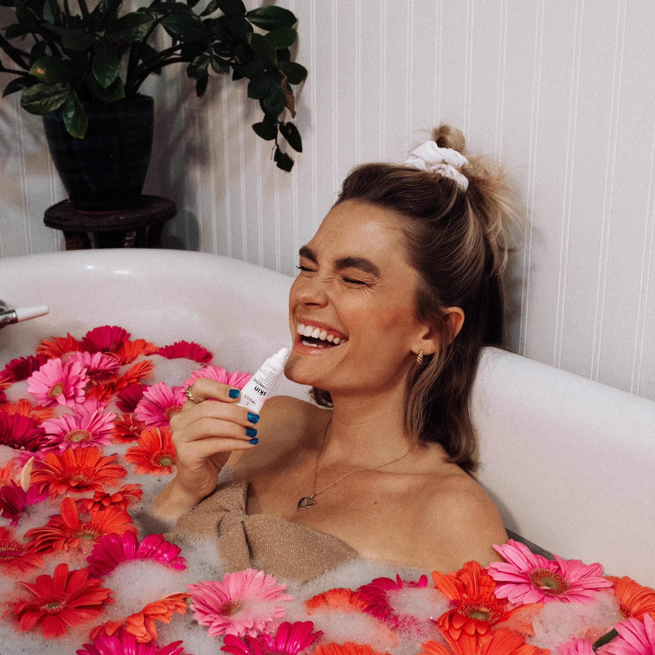 Woman in a bathtub filled with flowers, surrounded by a plant and white walls.