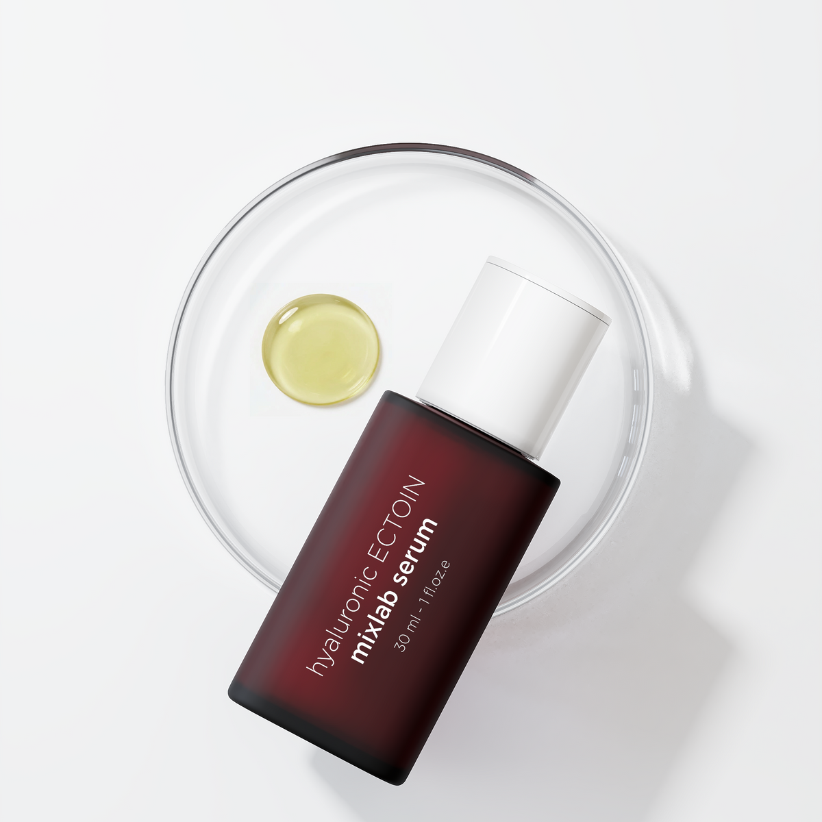 A bottle of miLAB serum with a dark reddish-brown body and white cap, placed diagonally on a clear glass dish containing a small gold droplet, on a clean white background.