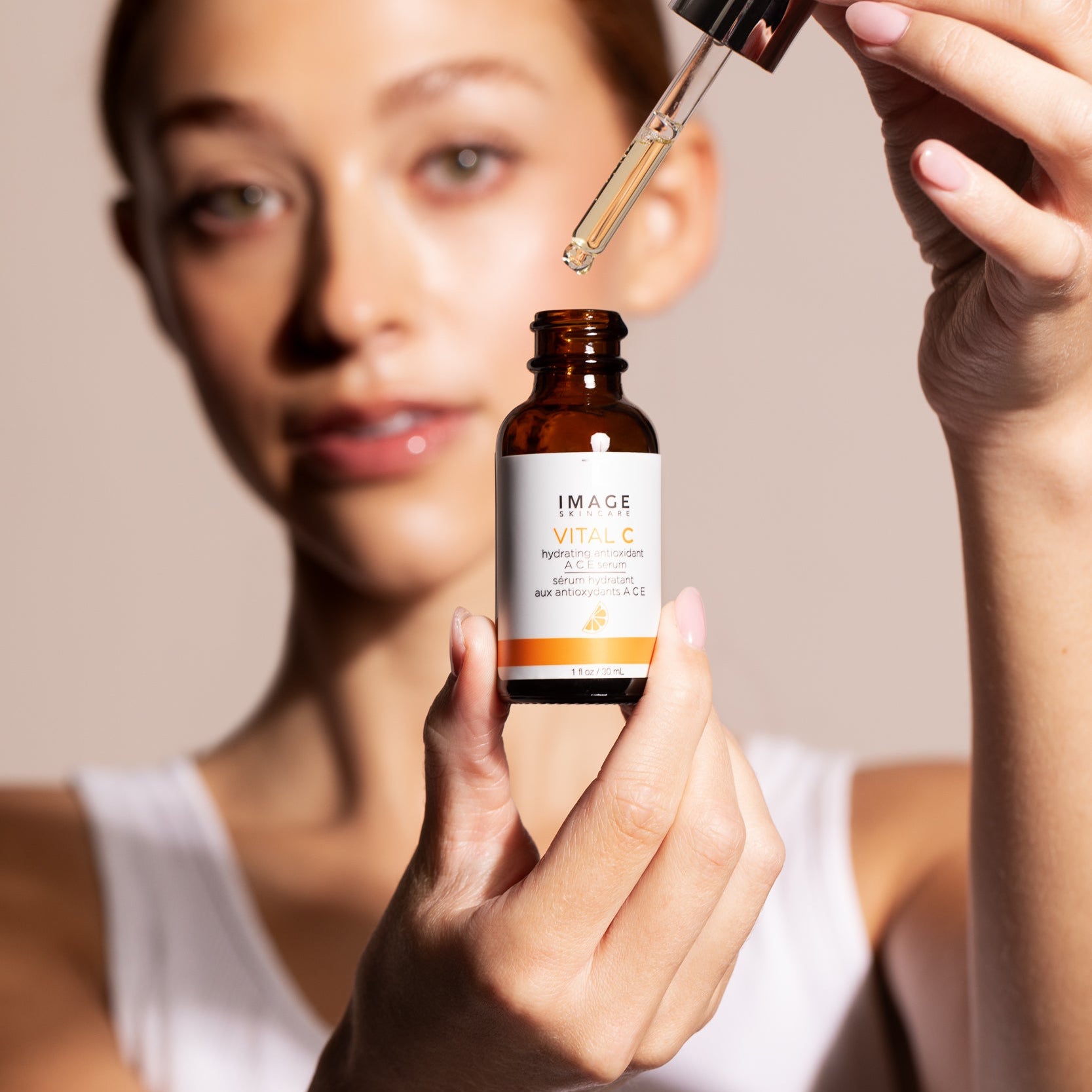 Woman holding a bottle of skincare product with a dropper above her face against a neutral background