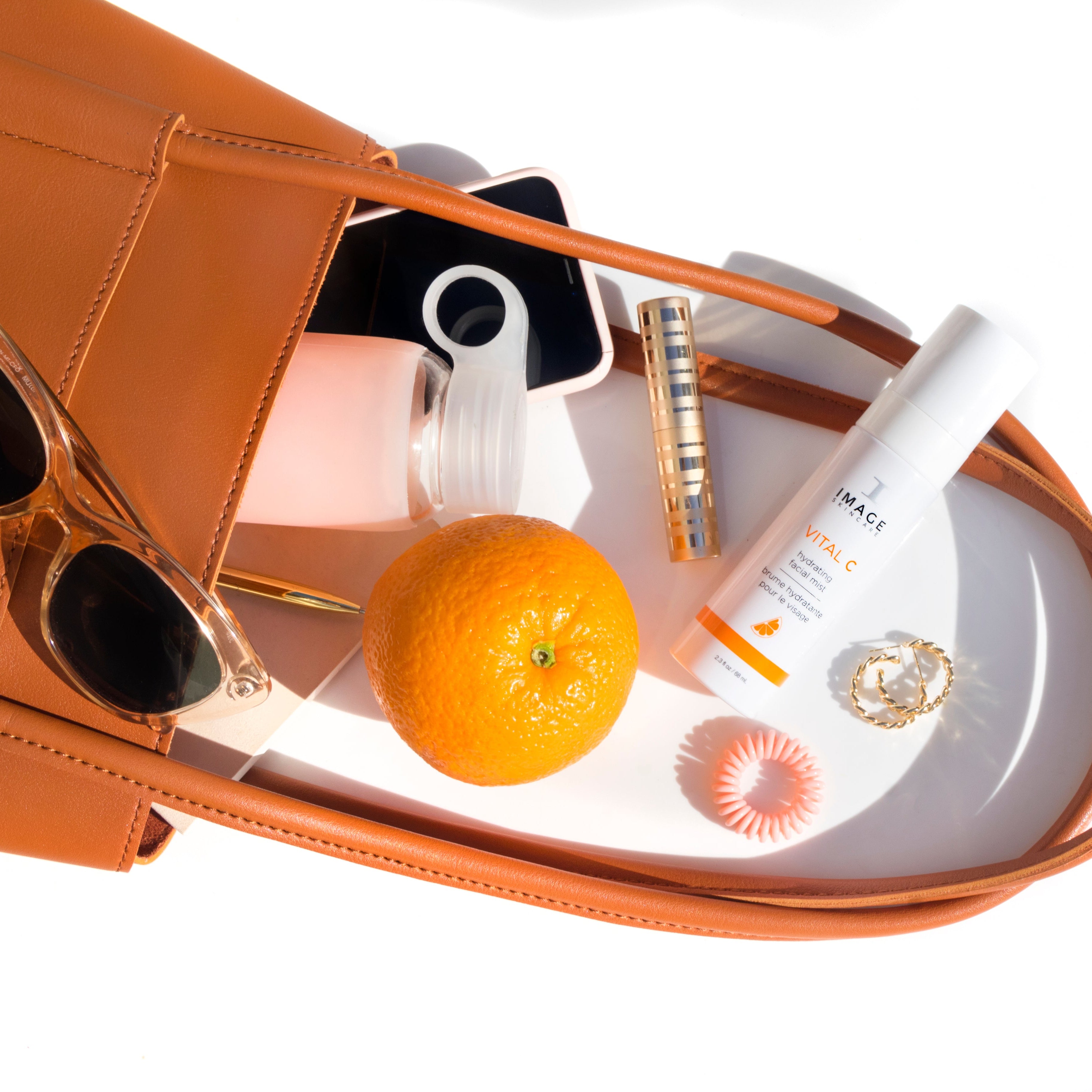 Brown leather bag with sunglasses, orange, and other items on a white surface