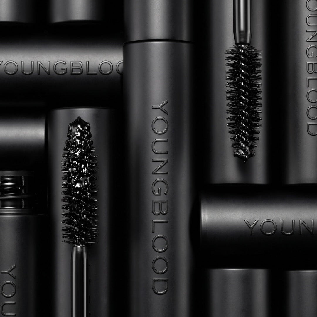Close-up of multiple YOUNGBLOOD mascara tubes arranged vertically, with two mascara wands shown upright against a dark black background.