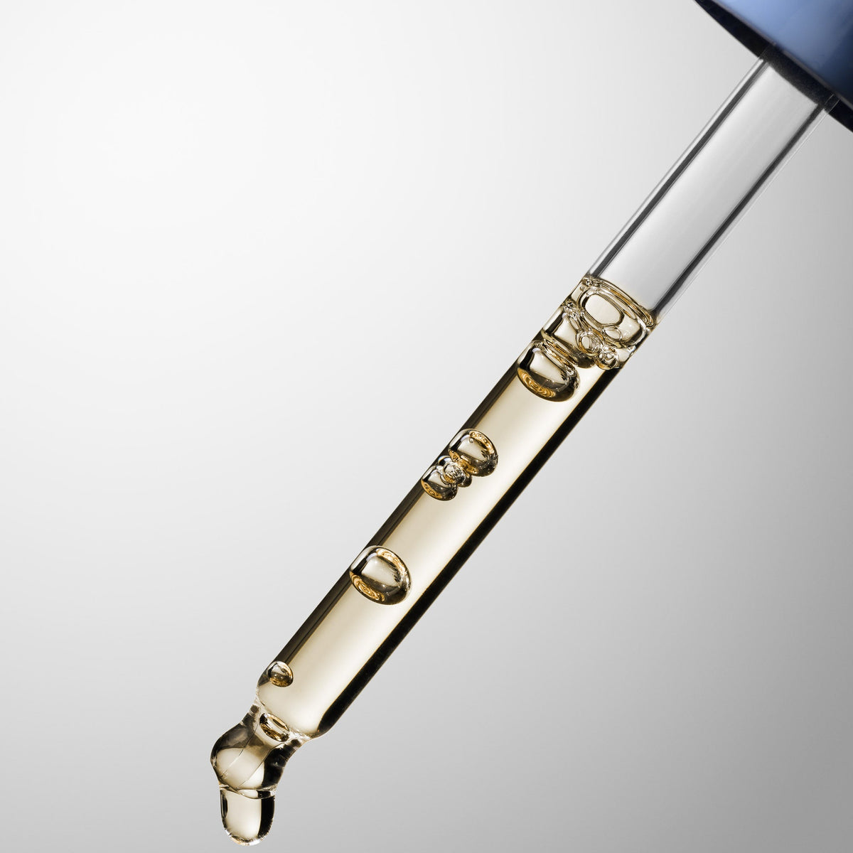 A close-up of HydroPeptides Syner-C+  glass dropper containing a transparent golden liquid, with one droplet falling below and a blue cap visible at the top against a light gray gradient background.